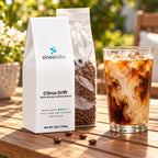 Citrus Drift Medium Roast Coffee, Whole Bean 12oz on a sunny patio table beside a tall glass of iced coffee with milk swirling in