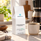 Vitality Mushroom Medium Roast Coffee, Ground 12oz on a kitchen counter beside a drip coffee maker with a full glass carafe and a mug of dark coffee